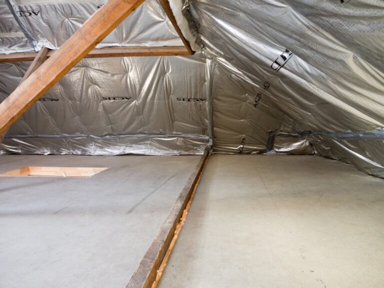 A worker in protective gear installing thick insulation batts in an unfinished attic.
