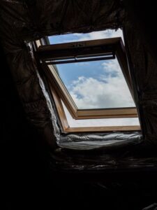 Homeowner examining attic insulation near roof framing while sunlight enters through a small access hatch